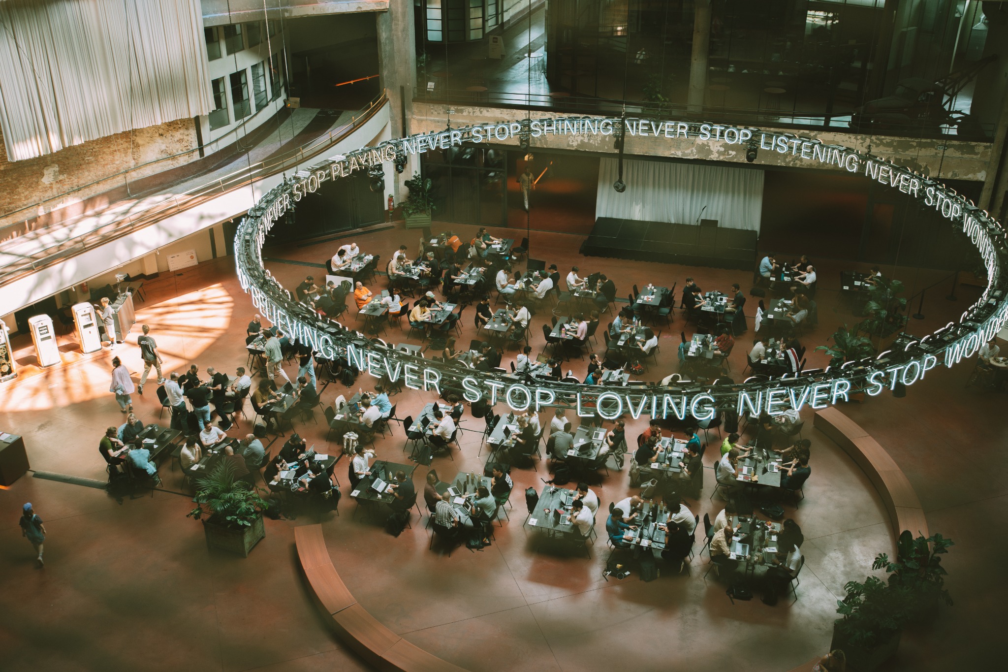 Dozens of builders working together at tables under a sunlit industrial ceiling during a community hackathon.